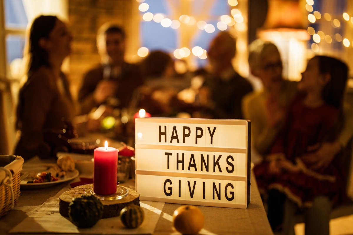 A group of people sitting around a table, embracing their thanksgiving traditions with a sign that says happy thanksgiving.