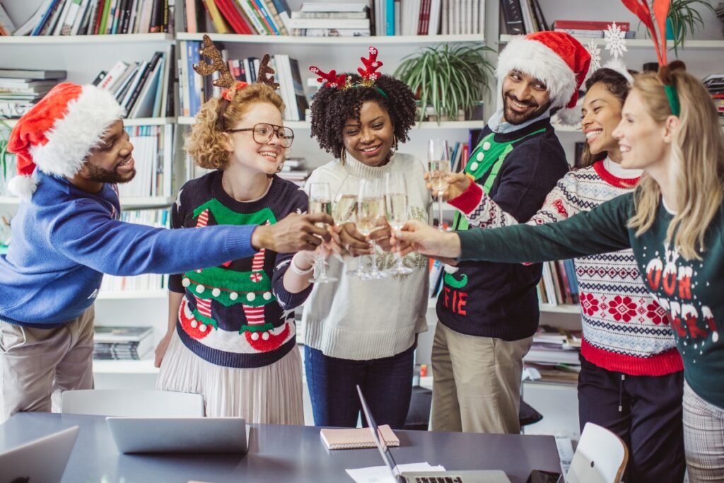 A group of people in Santa hats and ugly sweaters toasting in an office.