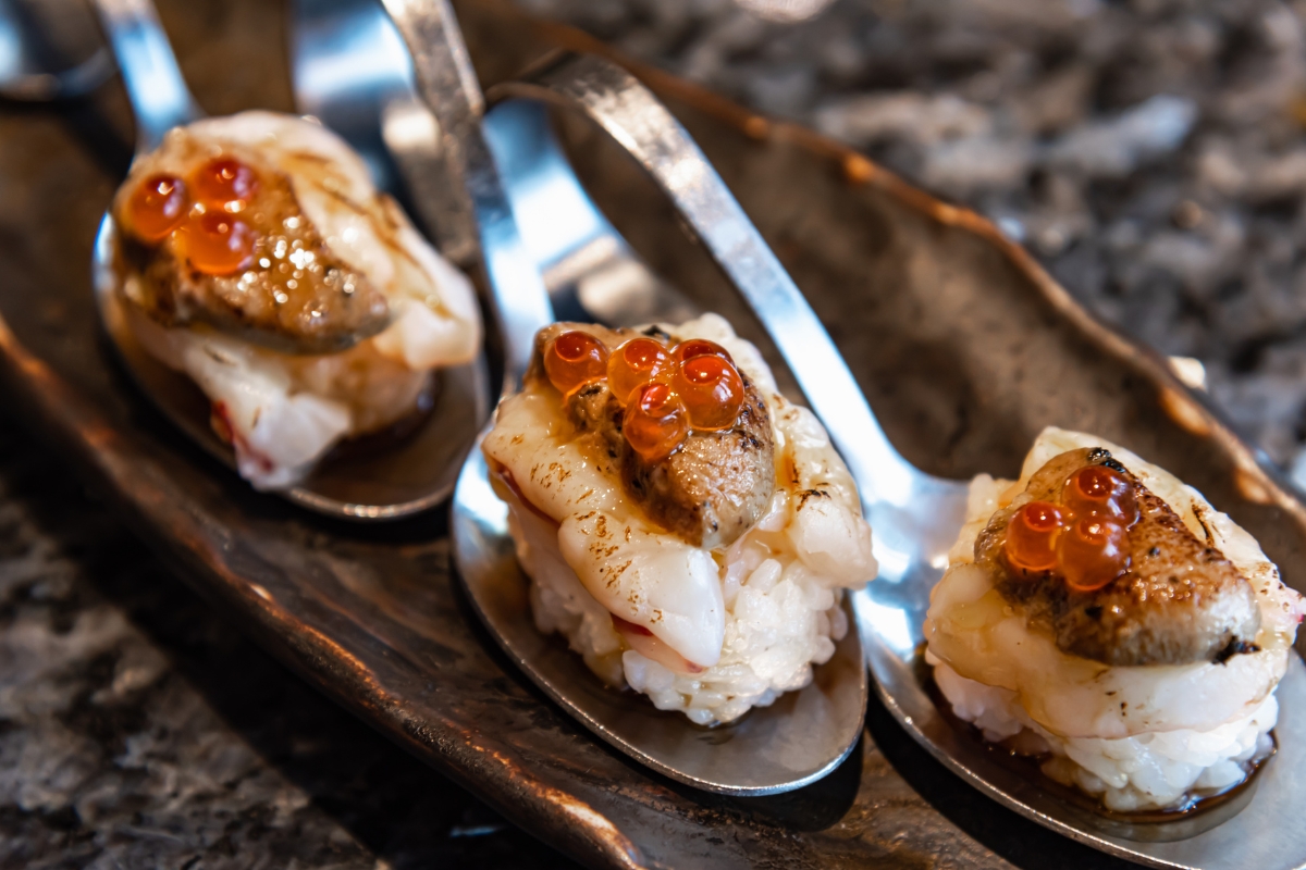 Three pieces of sushi with fish roe topping presented on individual metal spoons by a fusion cuisine catering service.