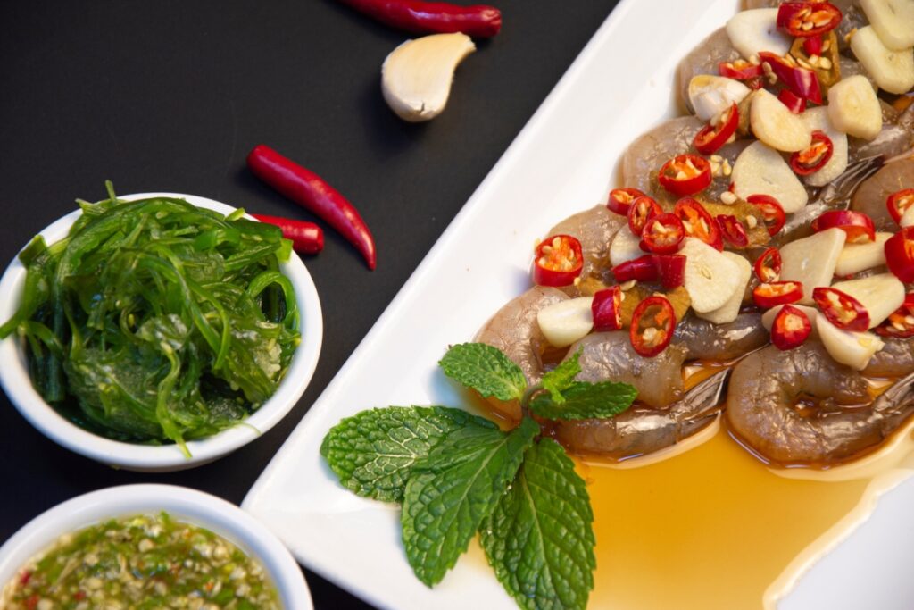 A plate of fusion cuisine featuring marinated fish with garlic and chili peppers, accompanied by a side of green herbs and a dipping sauce.