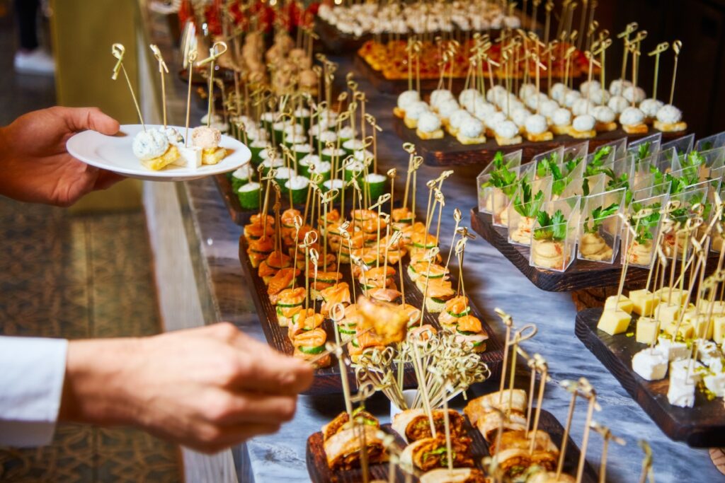 A person serves themselves appetizers from gourmet event food stations, featuring an array of skewered and plated bites.