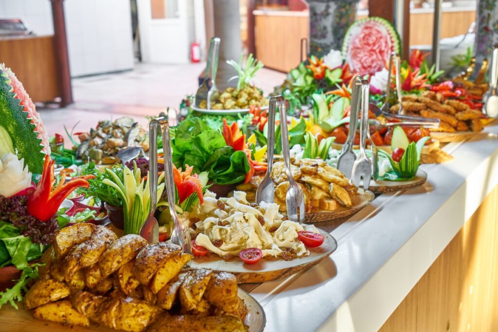 Buffet table showcasing a variety of dishes including salads, roasted potatoes, and fried fish, elegantly garnished at an event food station in a sunny venue.
