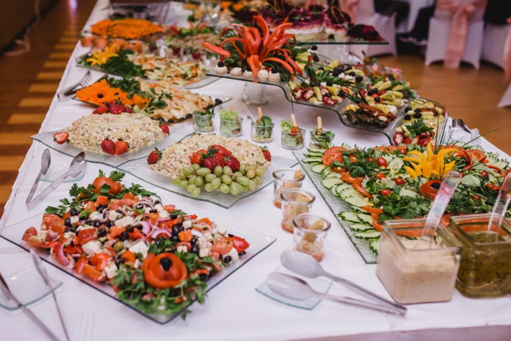 A buffet table at an event, laden with an array of salads, fresh fruits, and dressings at the food stations, garnished with vibrant edible flowers and decorative greens.