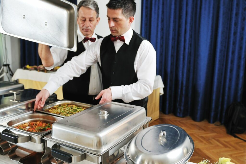 Two waiters in formal attire arranging and inspecting chafing dishes at interactive food stations at a catering event.