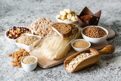 A variety of carbohydrate-rich foods displayed on a wooden board, including different types of pasta, rice cakes, bread, gluten-free catering options like grains and beans.