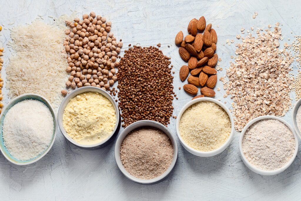 An assortment of grains and nuts, including rice, chickpeas, buckwheat, almonds, and oats arranged in rows, with various powders and flours in small bowls below them on a light background. Perfect for gluten-free catering.