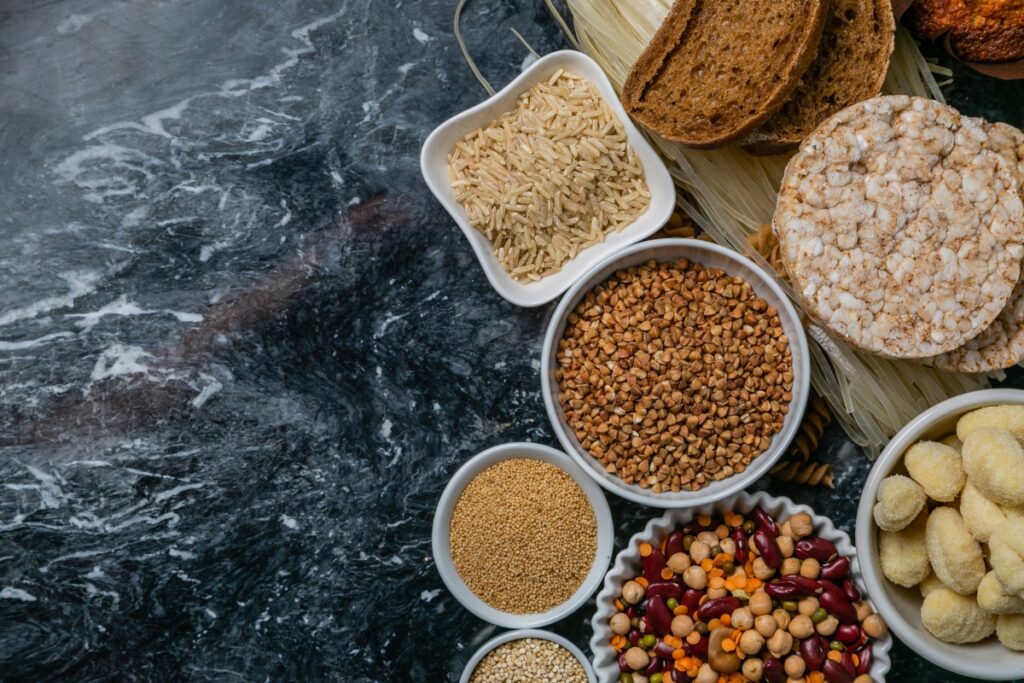 A variety of grains and bread displayed on a dark marble surface, including rice, buckwheat, millet, grains and seeds mix, pasta, bread slices, and rice cakes—all perfect for gluten free catering.