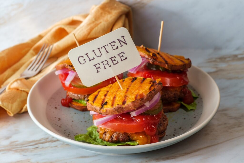 Three gluten-free burgers on a white plate, each adorned with lettuce, tomato, onions, and a toothpick displaying a "Gluten Free" sign. A mustard-colored napkin and a fork are in the background, epitomizing top-notch gluten-free catering.