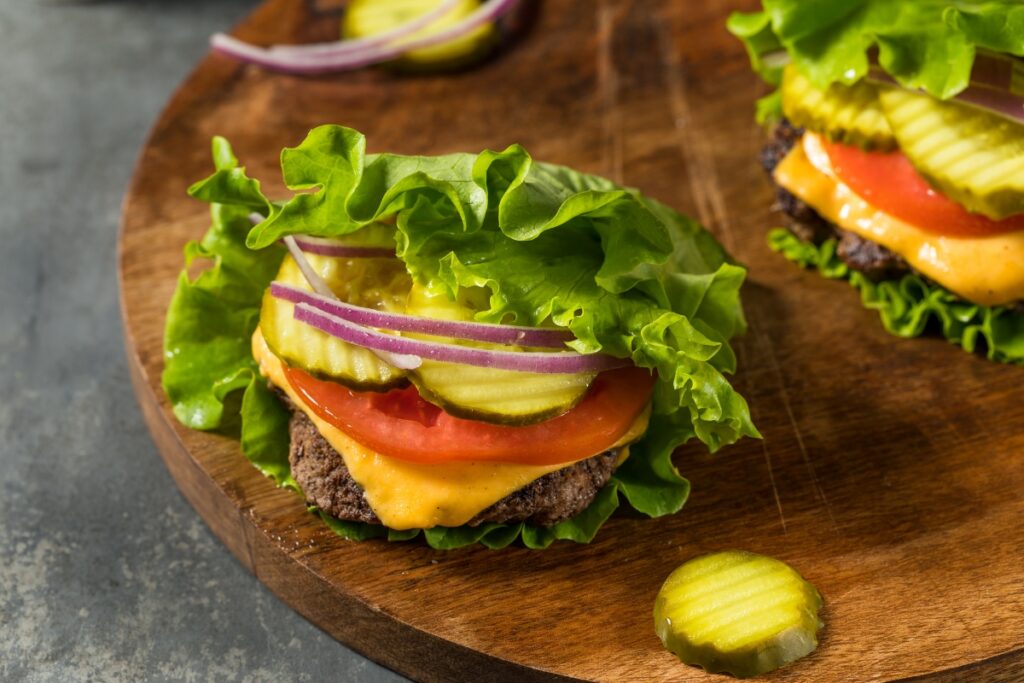 Two lettuce-wrapped burgers with cheese, pickles, tomato, and red onion are placed on a wooden surface. A single pickle slice rests beside the burgers. Perfect for gluten-free catering, these delicious bites offer all the flavor without the bun.