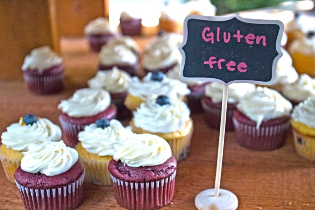 A display of gluten-free cupcakes with various colored frostings on a wooden counter. A small chalkboard sign in the foreground reads "gluten-free," perfect for your next gluten-free catering event.