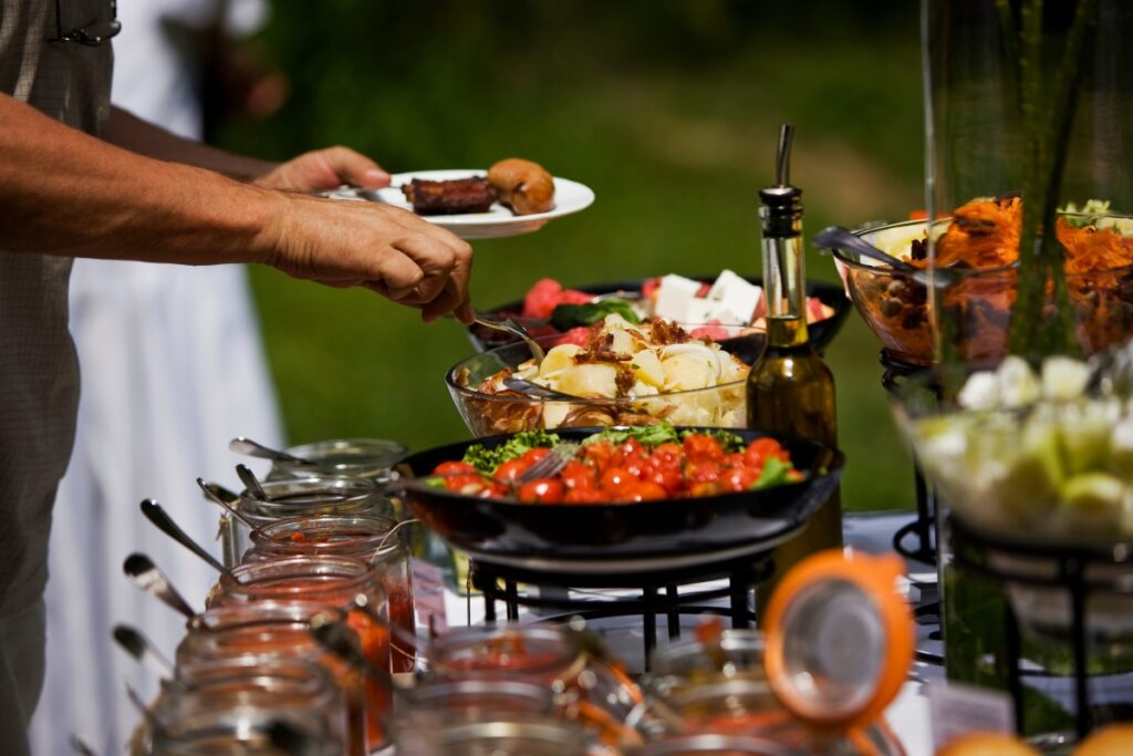 A person serving themselves food from a healthy catering buffet filled with various dishes, including salads and grilled items, outdoors.