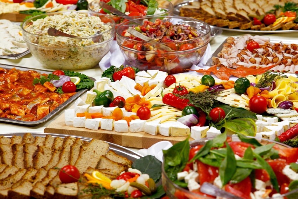 A buffet table spread with a variety of healthy dishes including cheeses, salads, sliced meats, and breads.