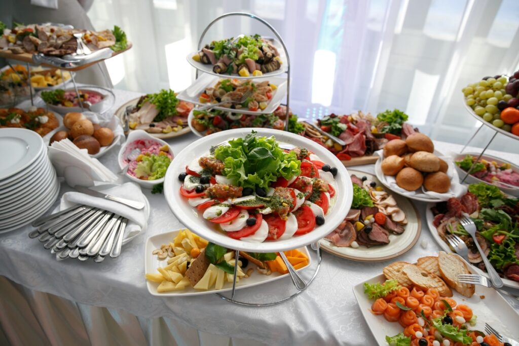 A buffet table spread with assorted healthy catering dishes including salads, meats, breads, and fruits, with plates and cutlery at the side.