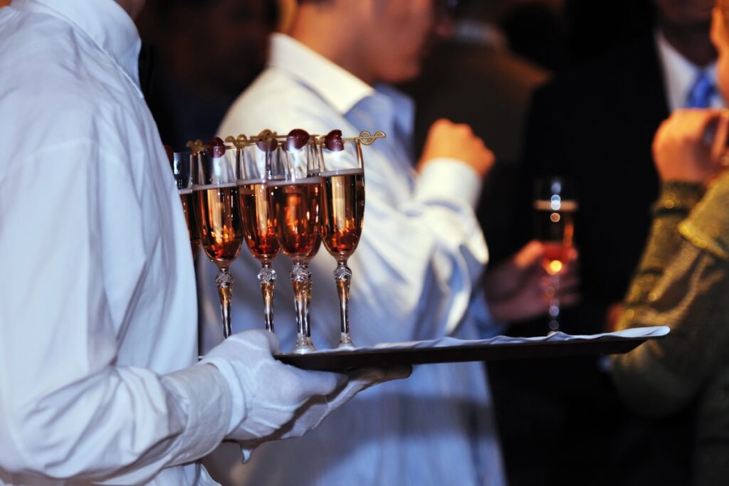 A server in a white uniform holds a tray with glasses of champagne garnished with cherries. People are blurred in the background, holding similar glasses of elegant mocktails.