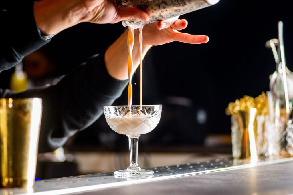 A bartender pours a creamy liquid from a shaker into a cocktail glass, expertly crafting one of the many enticing catering cocktails on offer, with bar tools and drinks visible on the counter.