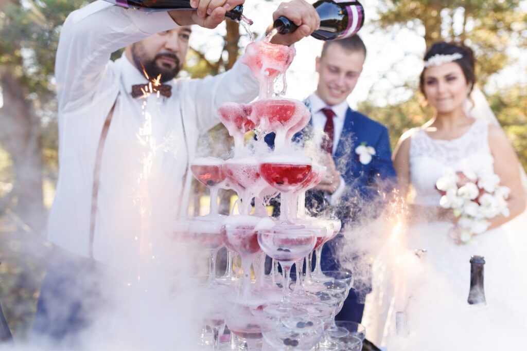 A man pours champagne into a pyramid of cocktail glasses at an outdoor event; a couple wearing wedding attire stands nearby, smiling. The catering team also offers a delightful selection of cocktails and mocktails for guests to enjoy.