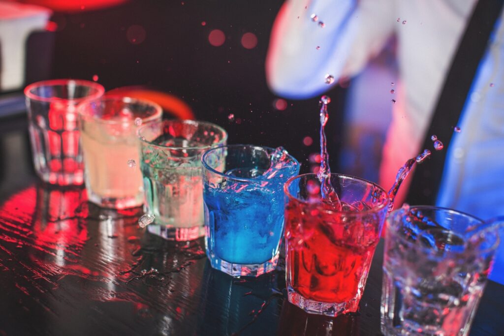 A row of six colorful catering cocktails spills over the edges of their glasses on a dark bar counter, with a person partially visible in the background.