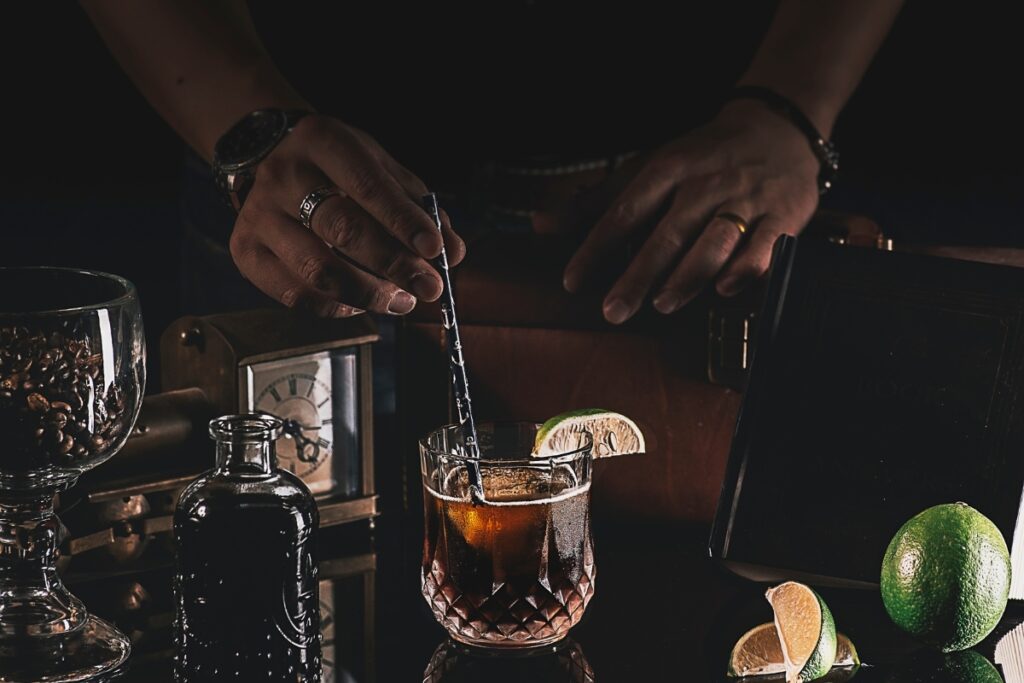 Close-up shot of a person stirring a mocktail in a glass with a straw, surrounded by a lime, a book, a clock, and a glass container with coffee beans, all set on a reflective surface.