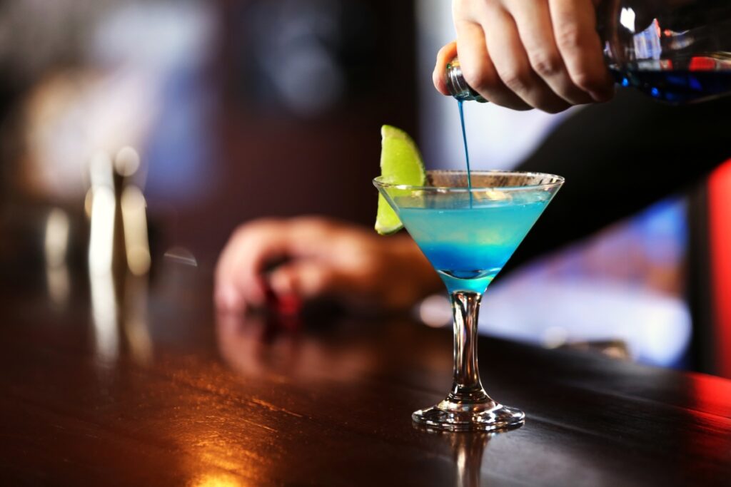 A bartender pours a blue cocktail into a martini glass garnished with a lime wedge on a wooden bar counter, showcasing their catering cocktails expertise.