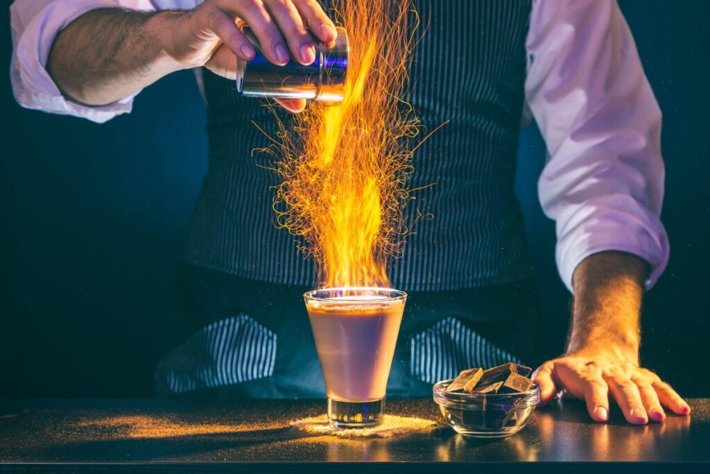 A bartender sprinkles powder into a flaming cocktail, creating a burst of sparks. A small bowl with wood chips is on the counter. The bartender, specializing in catering cocktails, is wearing a striped vest and white shirt.