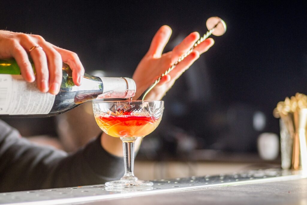 A person pouring a mocktail from a green bottle into a glass goblet, while stirring with a twisted bar spoon.