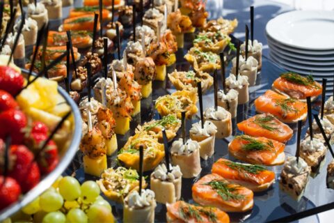 A buffet table with an assortment of finger foods and appetizers, including skewered shrimp, salmon with dill, fresh fruits, and various bite-sized snacks neatly arranged on a glass surface. For catering tips for large groups, ensure diversity in options to satisfy all tastes.