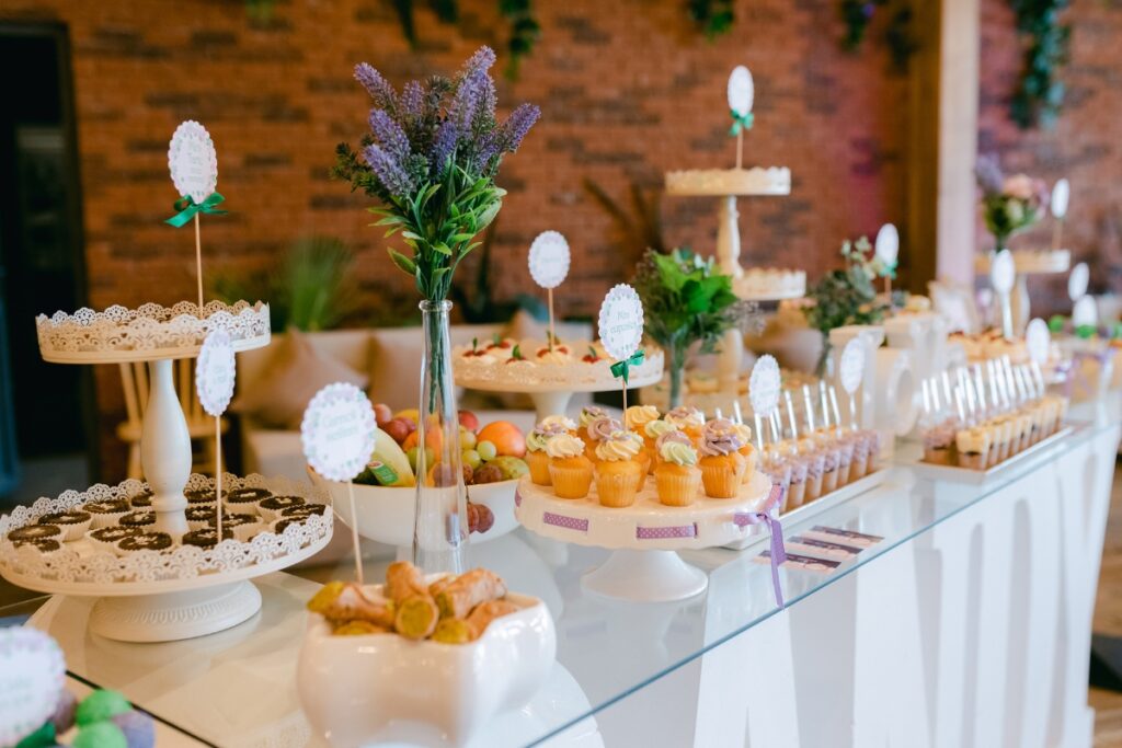 A dessert table is set with cupcakes, cookies, cake pops, and floral arrangements, displayed on various stands in a well-lit room with a brick wall backdrop—an ideal setup when considering catering tips for large groups.