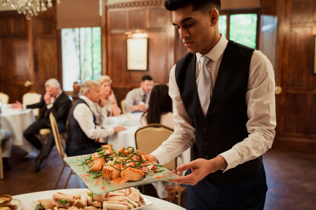 A server in a black vest and tie carries a platter of salmon and greens, showcasing excellent catering tips for large groups. People are seated at round tables in the background, engaged in conversation.