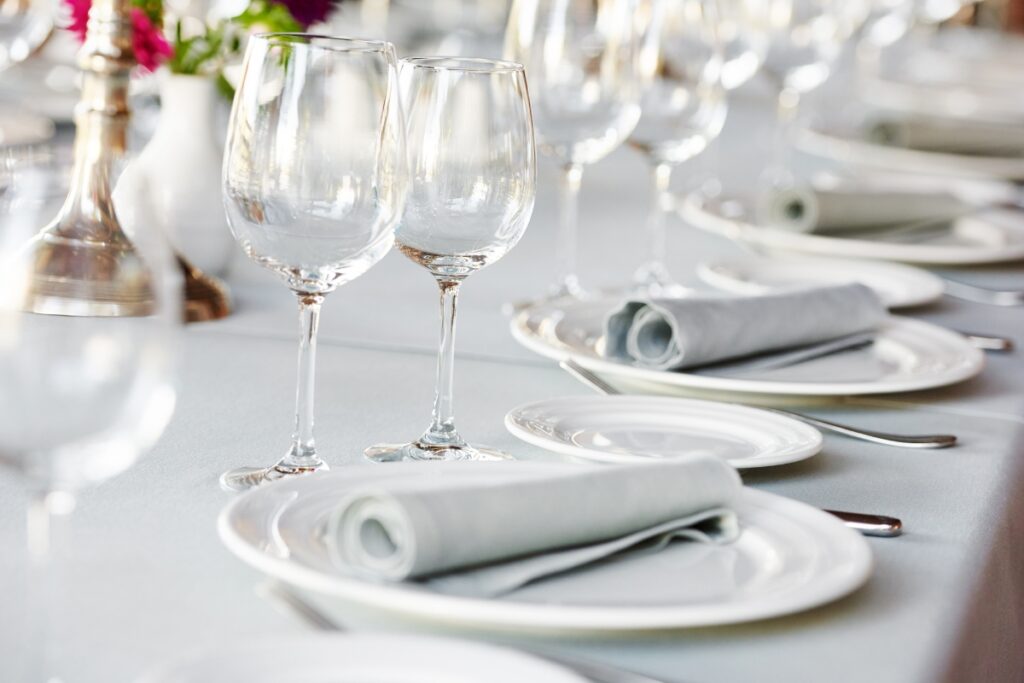 A neatly set dining table features white plates with folded gray napkins, clear wine glasses, and cutlery. Blurred flowers and a silver candlestick holder are in the background, offering an elegant touch—perfect for incorporating catering tips for large groups.