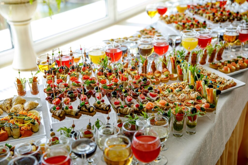 A large spread of assorted appetizers and drinks is displayed on a table near a window, featuring colorful finger foods, garnished treats, and various drinks in glasses. Perfectly executed catering tips for large groups are evident in every delectable detail.