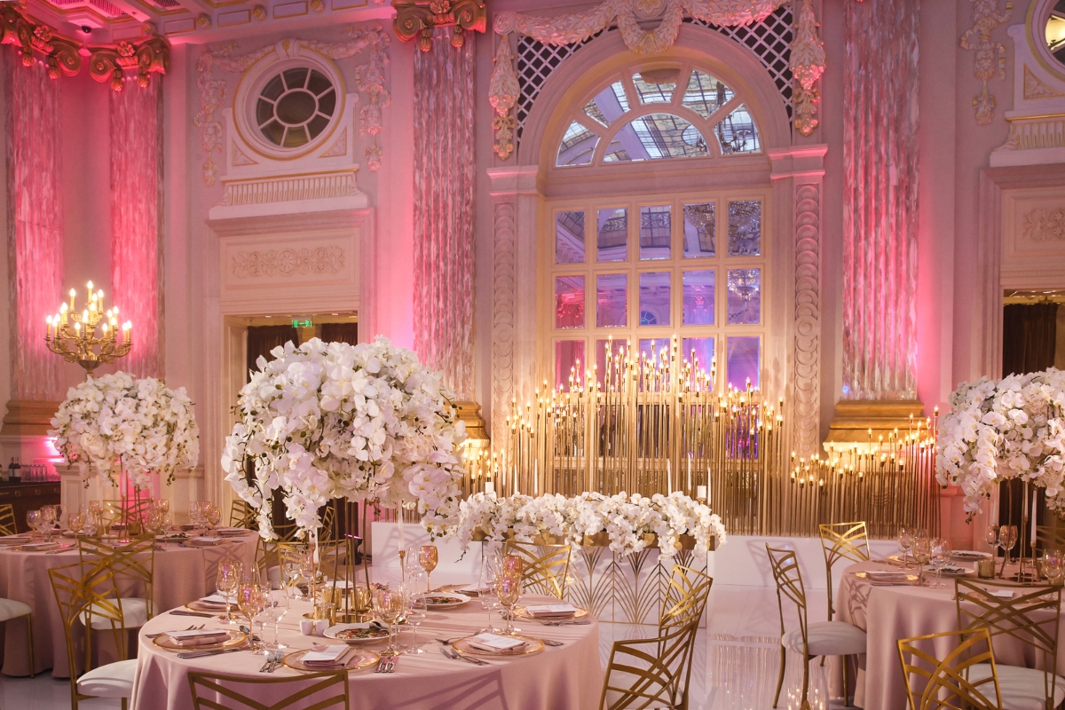 An opulent ballroom is adorned with lavish floral arrangements, gold-colored chairs, and round tables. Perfect for cultural weddings, the room features grand windows, ornate walls, and warm ambient lighting.