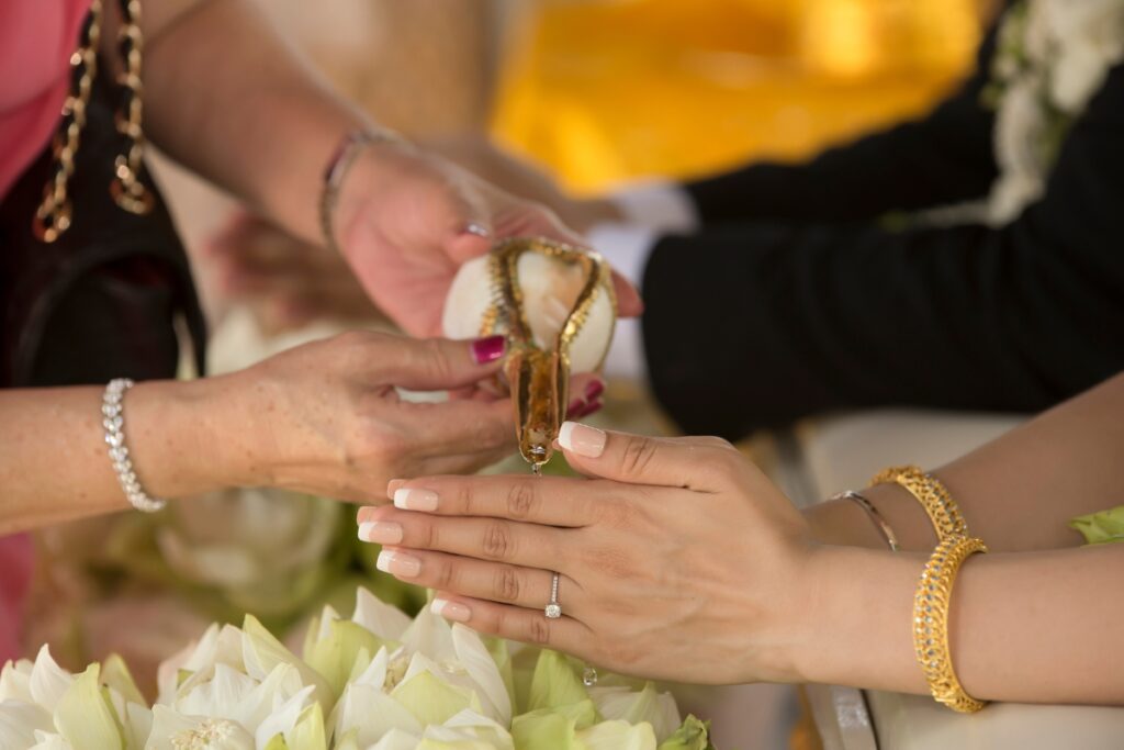 Close-up of hands in a ceremonial setting, with one hand pouring water over another's cupped hands. The recipients wear gold bracelets, and white flowers can be seen in the foreground—a beautiful moment often seen in cultural weddings.