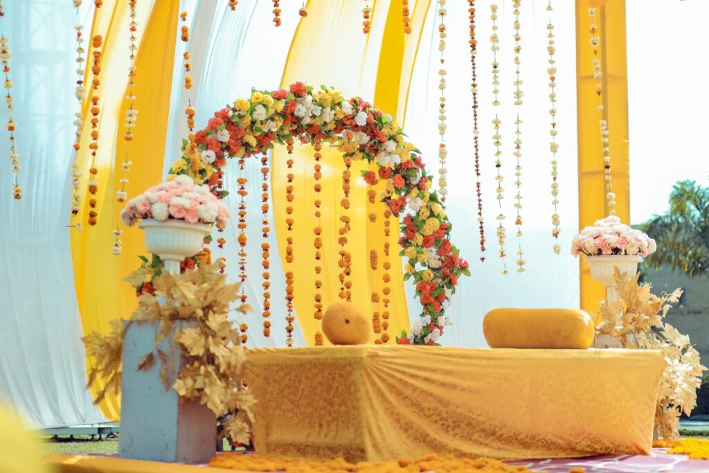 A decorative setup with a floral arch, yellow backdrop, and white vases with flower arrangements, perfect for cultural weddings or celebration events.