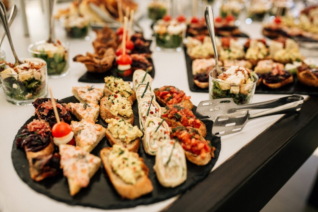 A variety of appetizers arranged on a black platter, including different types of bruschetta and small glass bowls with salads, perfect for cultural weddings, with serving utensils nearby.