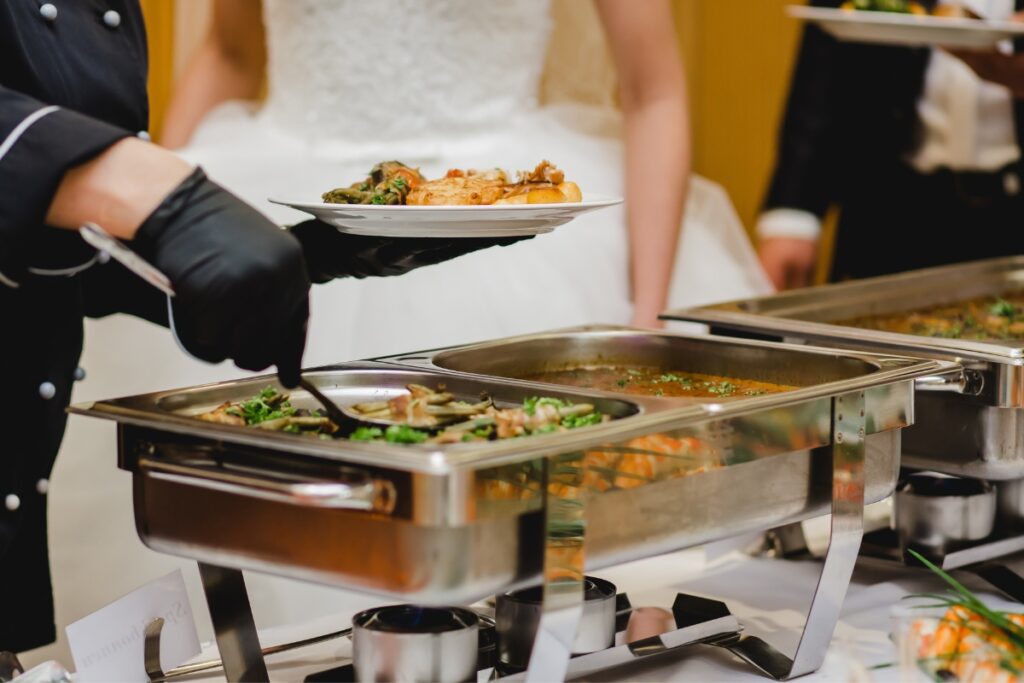 A person in a white dress is selecting food from a buffet spread at a cultural wedding, while someone in black gloves serves a dish. Various chafing dishes filled with delectable cuisine are visible.