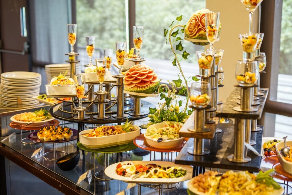 A buffet table with various dishes and appetizers, including salads in glasses and a decorative fruit arrangement, reminiscent of cultural weddings, arranged on multiple levels. Plates are stacked to the side.