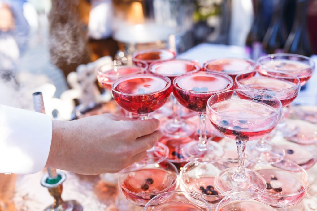 Hand reaching for a glass from a tower of pink cocktails garnished with berries, with steam visible nearby, resembling an elegant setup often seen at cultural weddings.