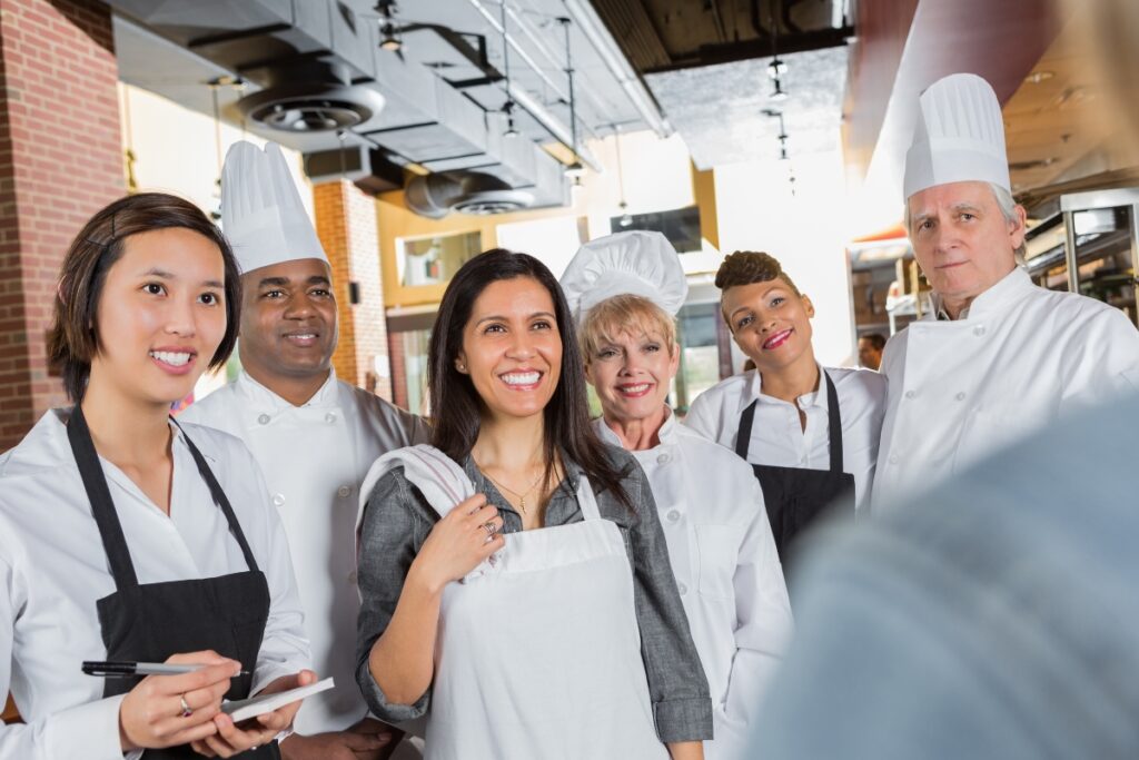 A group of chefs and a woman wearing an apron, possibly preparing for one of the many delicious dishes often found at cultural weddings, stand together in a kitchen. Everyone is smiling and looking at the camera.