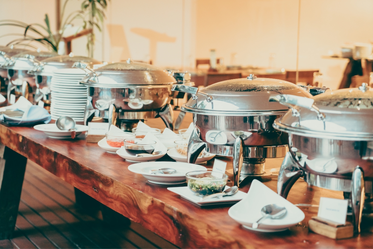 A buffet table with several chafing dishes, plates, bowls, and various food items arranged neatly on a wooden surface showcases our custom catering services.