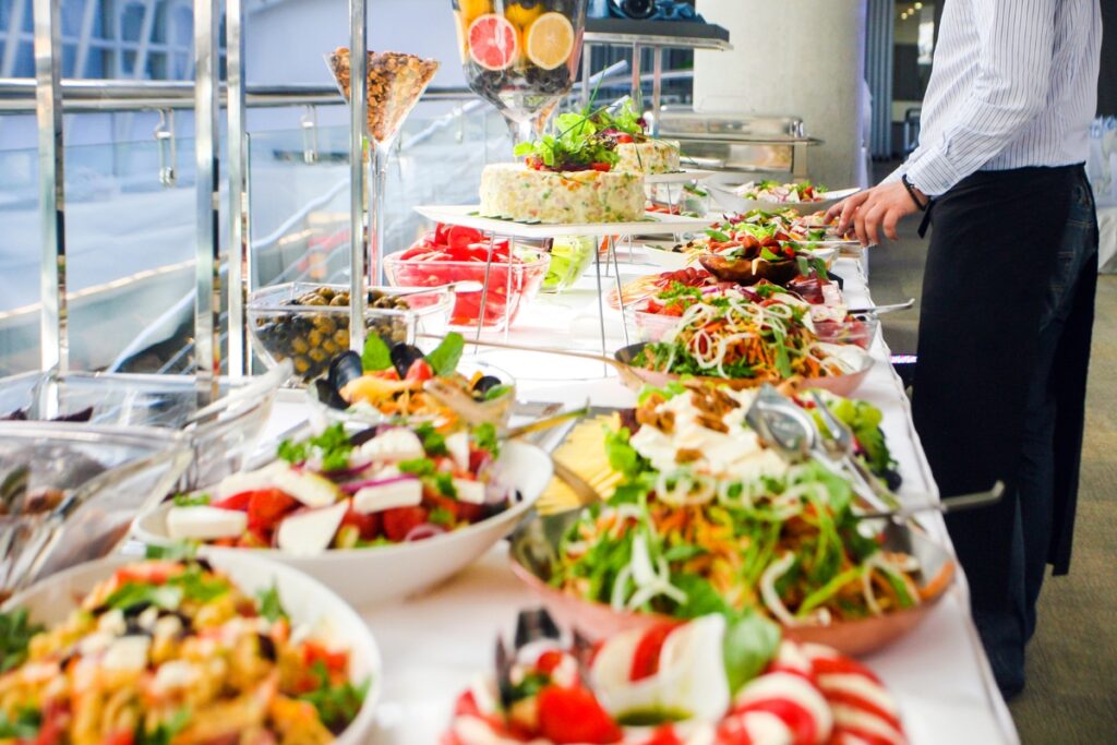 A person serves food from a buffet table laden with various salads, pasta dishes, fruit, and desserts, meticulously arranged by custom catering services.