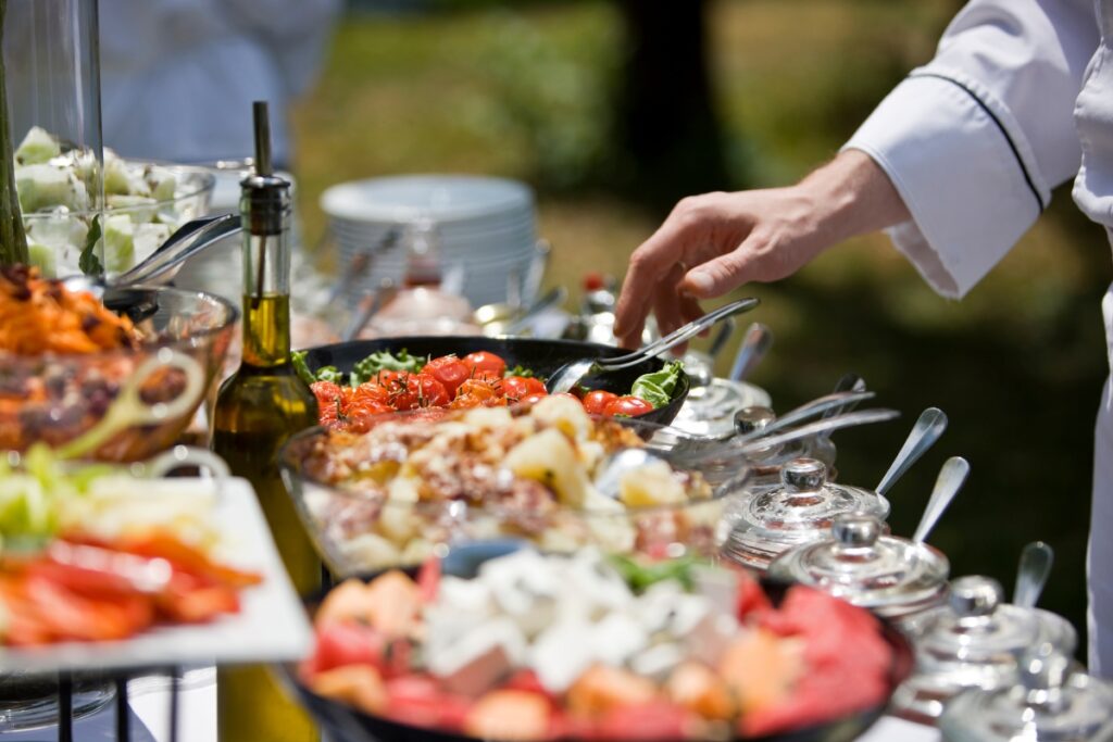 A person in a chef's uniform is serving food from a buffet table with various dishes, including vegetables, salad, and an olive oil bottle, under natural daylight. It's an exquisite display of custom catering services tailored to perfection.