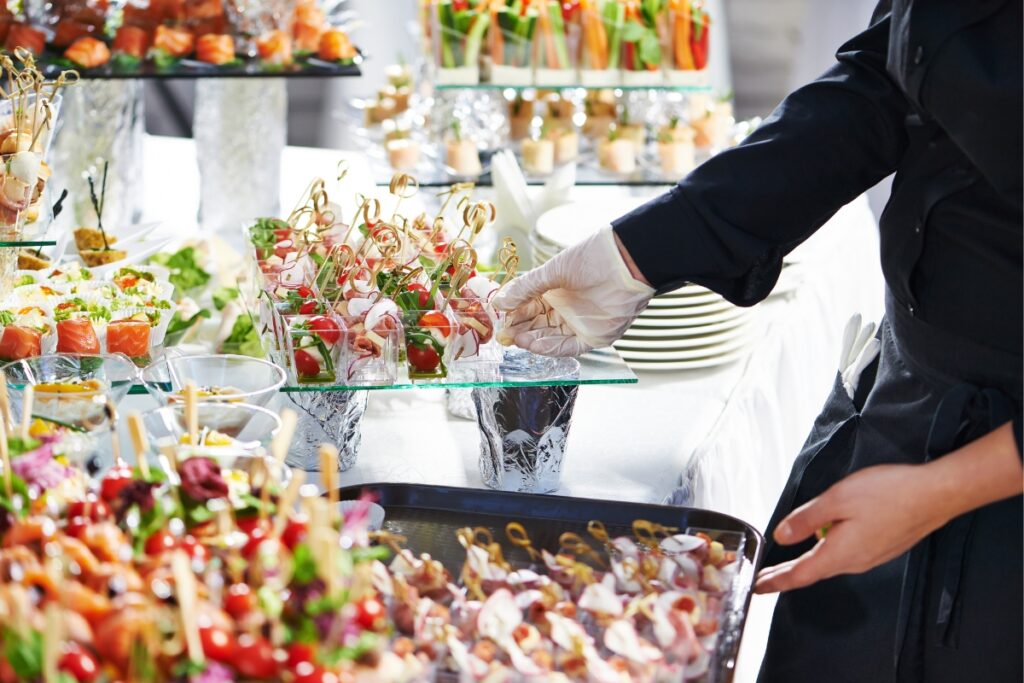 A person wearing a black uniform and a glove serves an assortment of appetizers on skewers and in small cups, displayed on a buffet table at an event, showcasing the elegance of custom catering services.