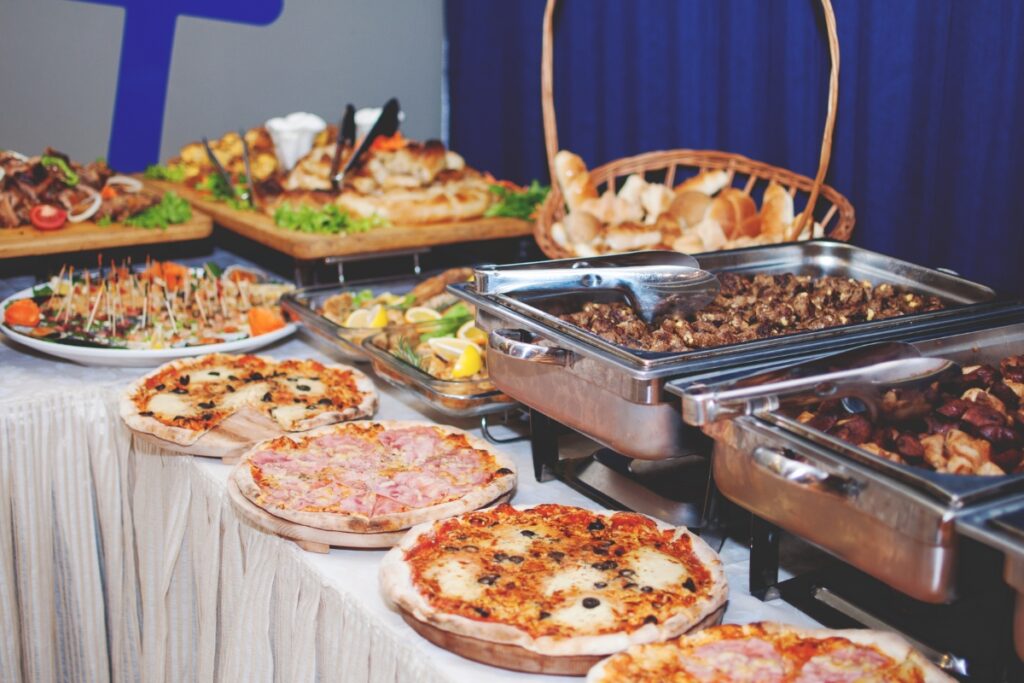 A buffet table with various dishes, including pizzas, rolls, meats in chafing dishes, skewers, and assorted appetizers, showcases our custom catering services.