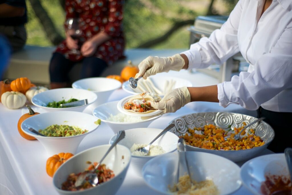 A person serving food into a taco shell from various bowls containing toppings on a buffet table decorated with small pumpkins, showcasing custom catering services.