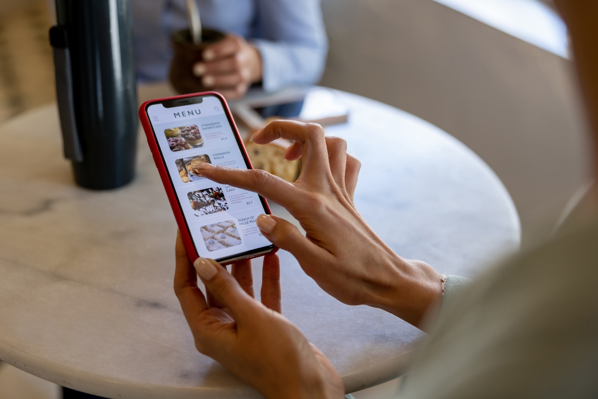 A person browsing a menu on a smartphone, utilizing catering technology with a touch of their finger. Another individual is seen partially in the background sitting at a table.