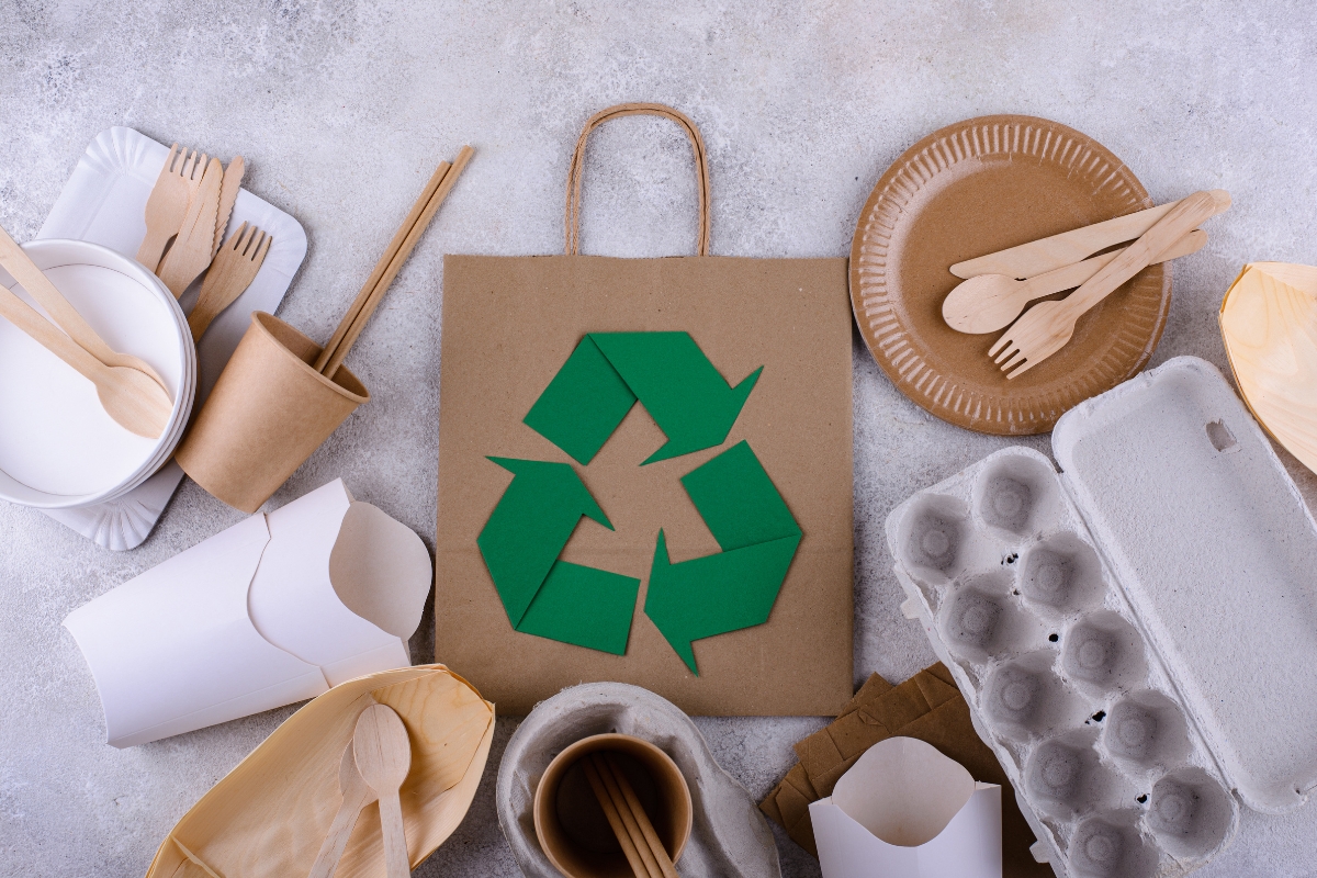 A recycling symbol on a paper bag, surrounded by various eco-friendly items including disposable plates, cups, cutlery, an egg carton, and containers—all made from paper or biodegradable materials—highlights the integration of catering technology in sustainable practices.