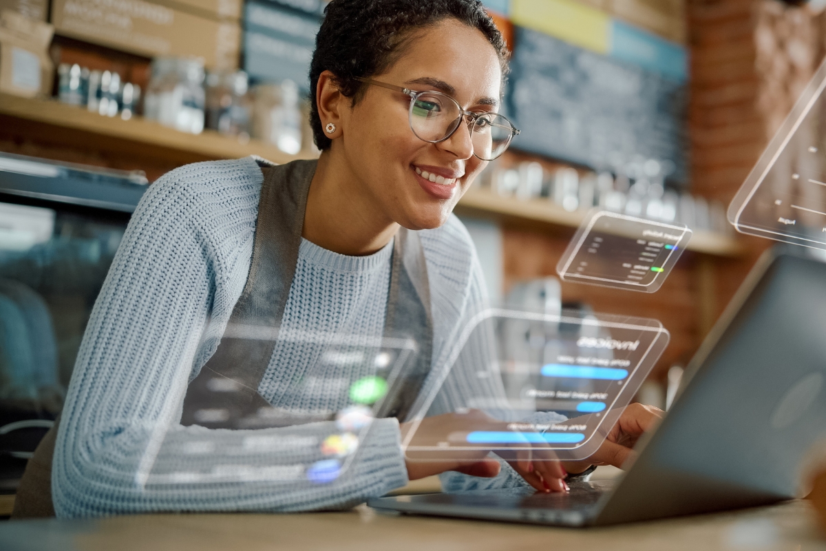 A person wearing glasses and a light blue sweater is using a laptop in a modern cafe with floating digital interface graphics around them, exemplifying the seamless integration of catering technology.