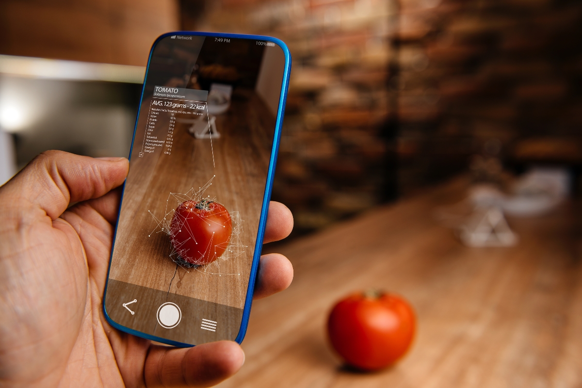 A person holds a smartphone using cutting-edge catering technology augmented reality to analyze a tomato on a wooden table, displaying nutritional information on the screen.