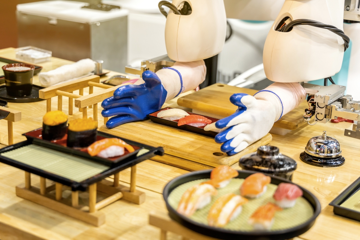 A robot with blue gloves makes sushi at a wooden table, showcasing the latest in catering technology, surrounded by various sushi dishes and a small bell.