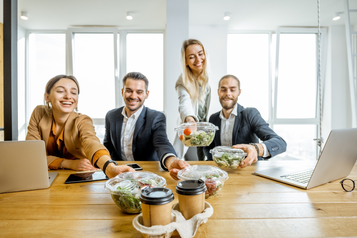 Four colleagues in business attire enjoy a lunch break in a bright office, sharing salads and coffee at a wooden table equipped with laptops and a tablet, thanks to convenient office catering solutions.